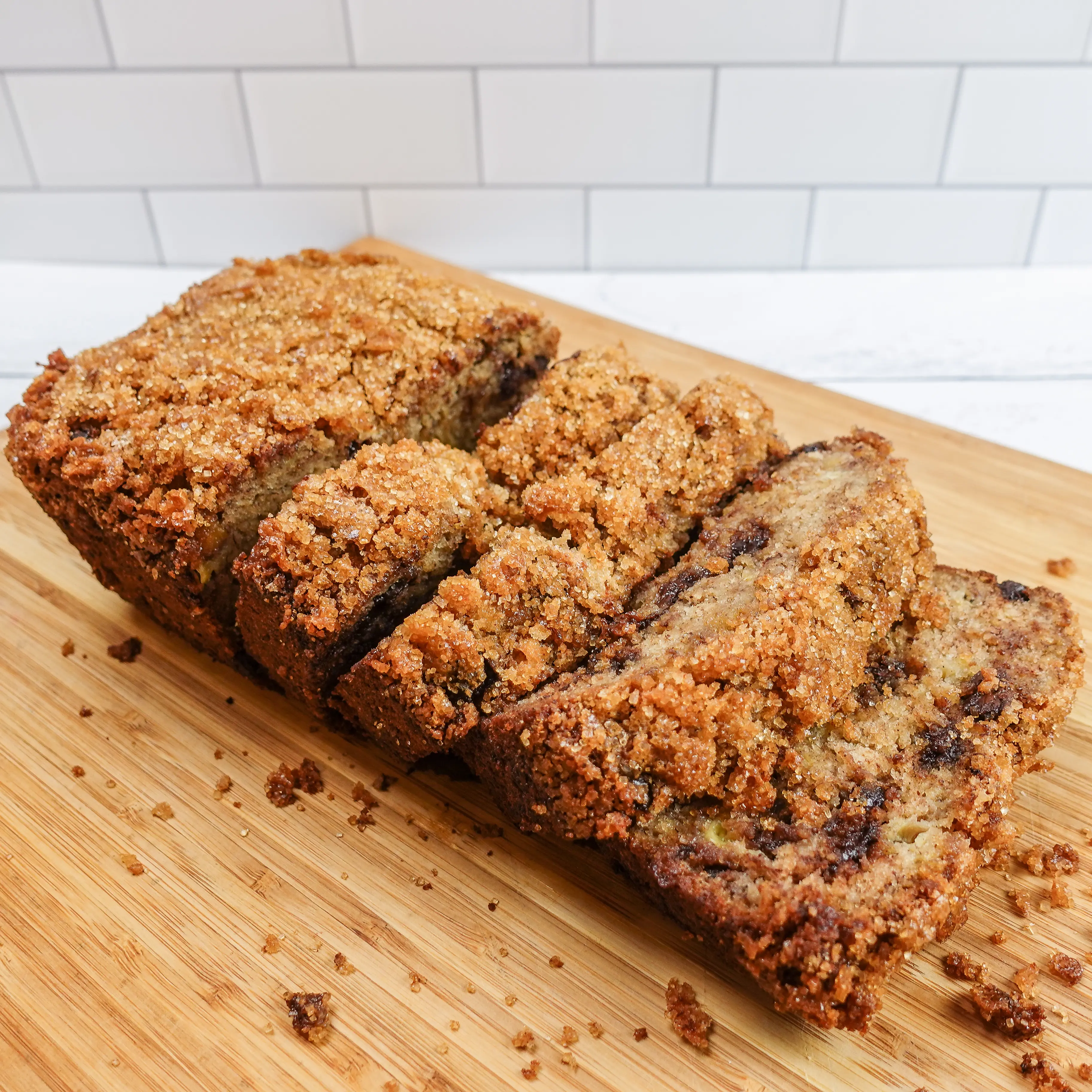 Warm, crumbly, delicious banana bread with melting chocolate chips inside, cut into slices on a wooden cutting board surrounded by crumbs, on a white wood plank counter with a white tile backsplash.