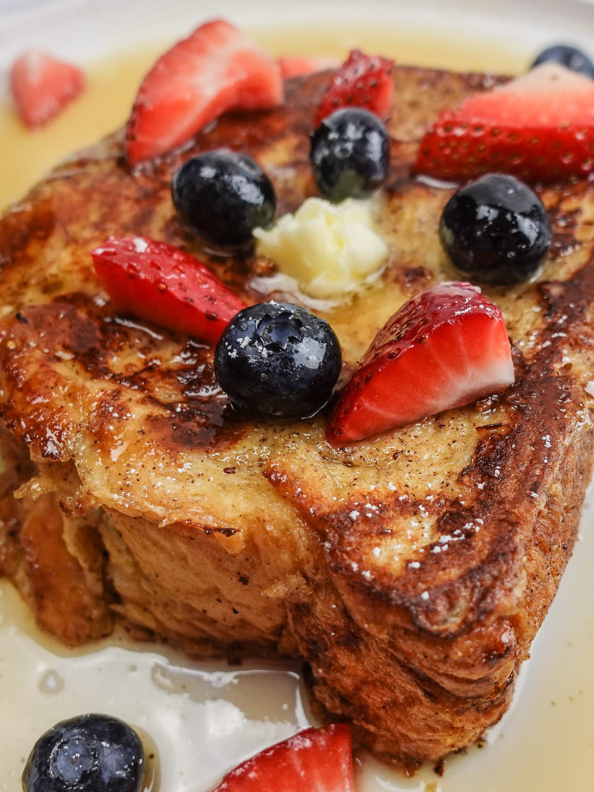 French Toast slice on a white plate, topped with butter, powdered sugar, sliced strawberries, blueberries, and maple syrup.
