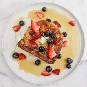 Overhead view of a French Toast slice on a white plate, topped with butter, powdered sugar, sliced strawberries, blueberries, and maple syrup, on a white marble countertop.