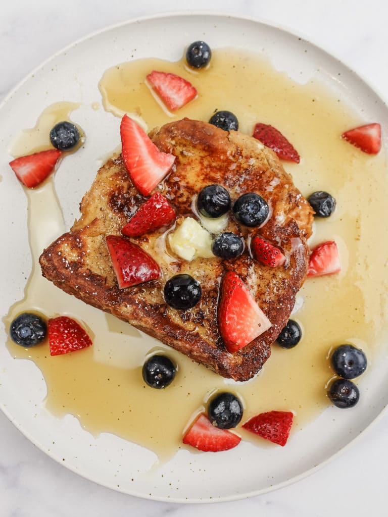 French Toast slice on a white plate, topped with butter, powdered sugar, sliced strawberries, blueberries, and maple syrup. 