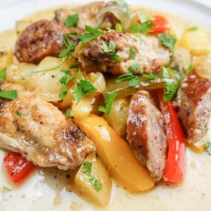 Closeup view of Italian chicken scarpariello including chicken wings, italian sausage, bell peppers, potatoes, banana peppers, and chopped italian parsley on a white ceramic dish.