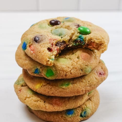 Soft and chewy M&M cookies stacked on top of one another, on a white marble countertop with a white tile back splash.
