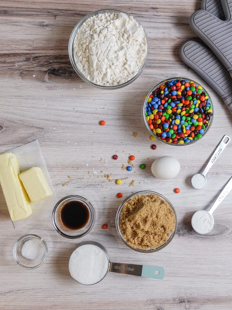 M&M Cookie Ingredients including all-purpose flour, M&M candies, an egg, baking soda, corn starch, salt, light brown sugar, vanilla extract, granulated sugar, and butter organized on a light grey and brown wooden counter next to grey oven mits.