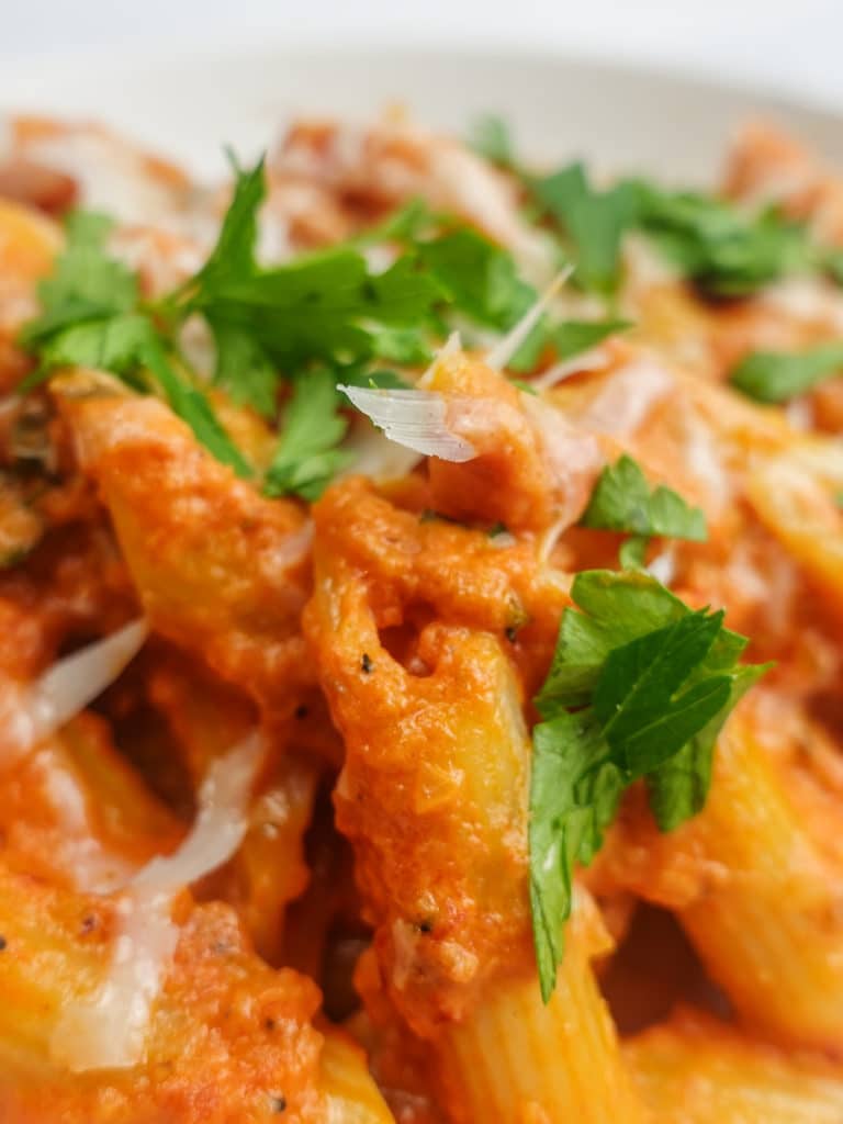 Closeup view of very thick and creamy Penne Alla Vodka pasta dish on a white plate garnished with grated pecorino romano cheese and chopped italian parsley.