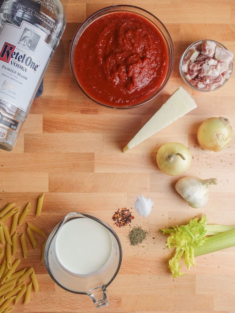 Penne Alla Vodka recipe ingredients including vodka, crushed tomatoes, pancetta, pecorino cheese wedge, onions, garlic, celery, cream, and penne pasta, organized on a wood chopping block countertop.