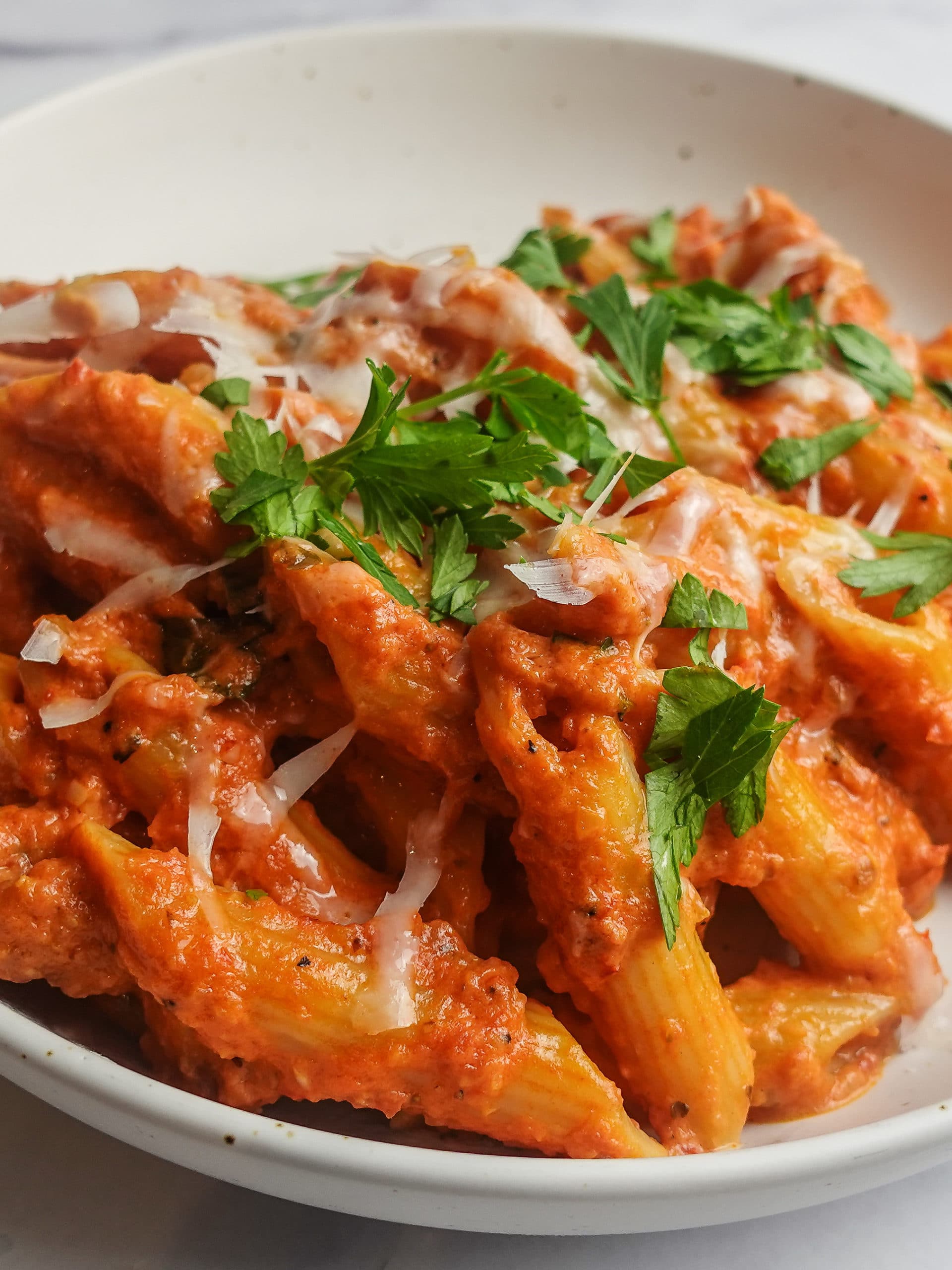 Very thick and creamy Penne Alla Vodka pasta dish on a white plate garnished with grated pecorino romano cheese and chopped italian parsley.