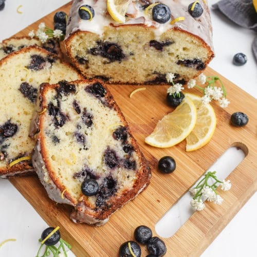 Overhead view of a sliced loaf of lemon blueberry bread with a white frosting, on a small wooden cutting board, decorated with lemon slices, blueberries and white flowers.