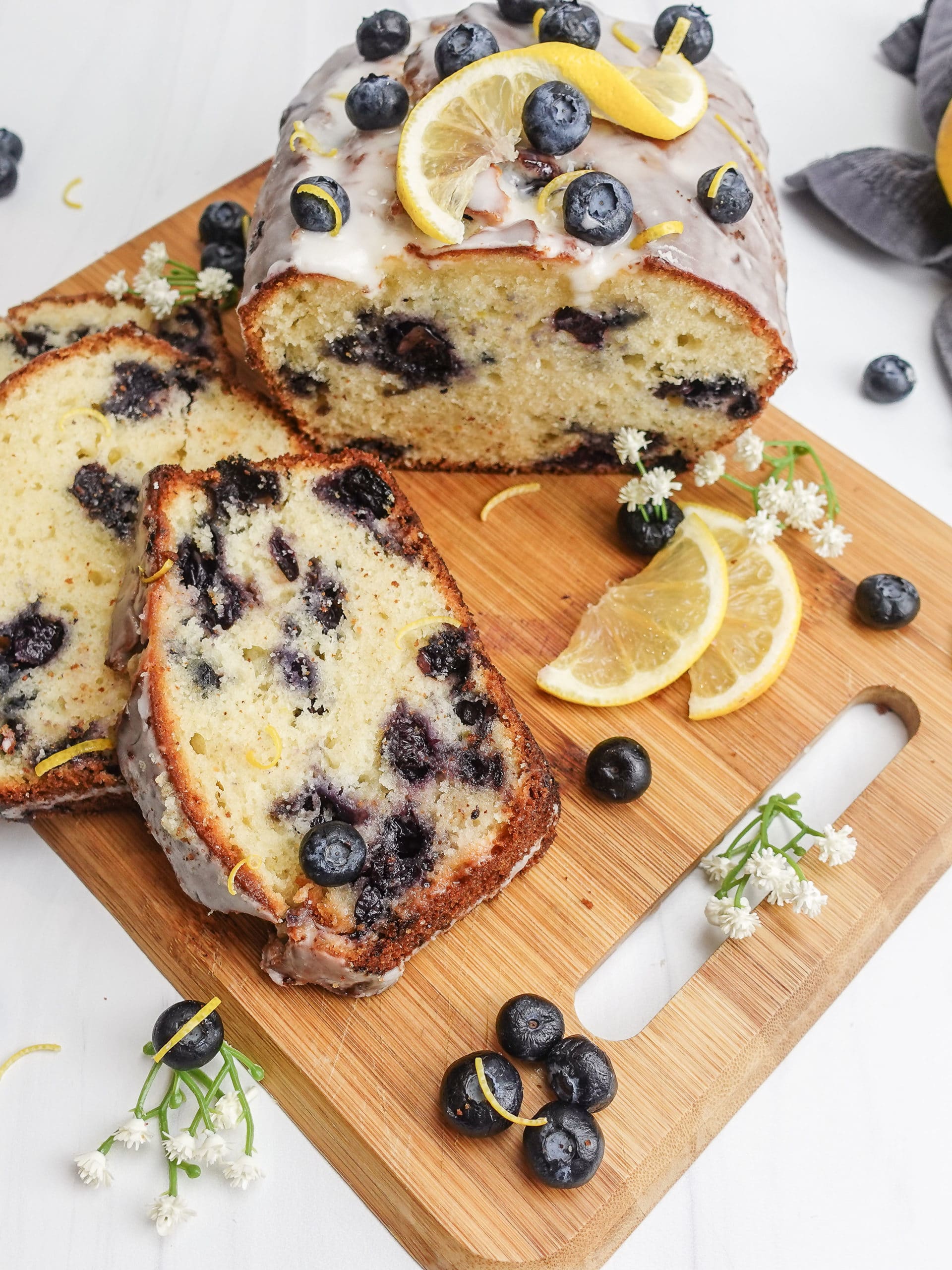 Overhead view of a sliced loaf of lemon blueberry bread with a white frosting, on a small wooden cutting board, decorated with lemon slices, blueberries and white flowers.