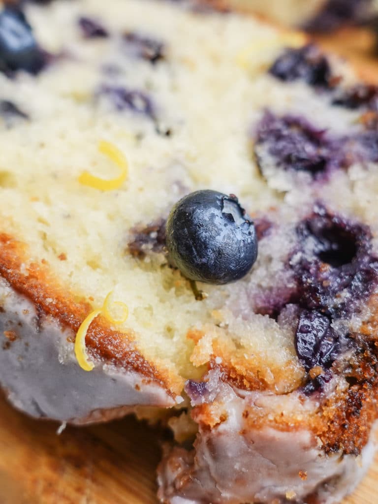 Closeup view of a slice of lemon blueberry bread with a frosting coating, garnished with blueberries and lemon zest.