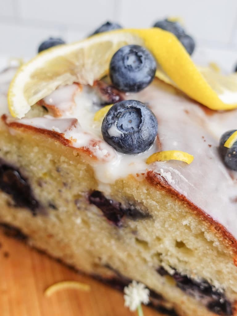 Overhead view of a loaf of lemon blueberry bread with a white frosting, on a small wooden cutting board, decorated with lemon slices, blueberries.