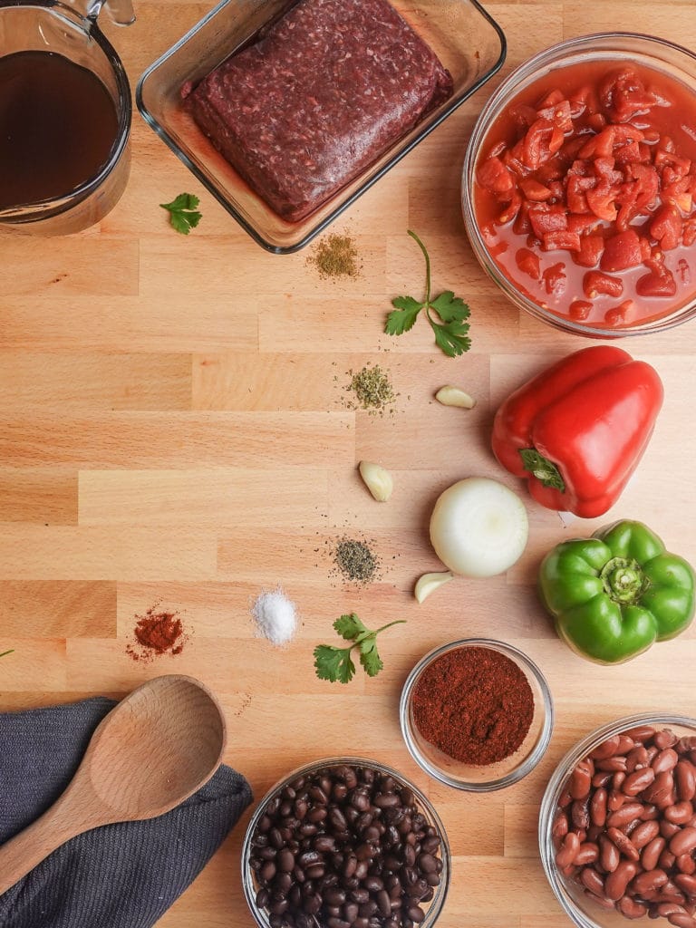 Simple and delicious chili recipe ingredients including beef broth, ground beef, diced tomatoes, bell peppers, onion, garlic, beans, and seasonings laid out on a wood chopping block counter, next to a wooden spoon on a grey kitchen towel.